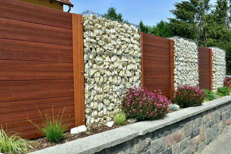 Moderner Sichtschutzzaun, abwechselnd aus mit Naturstein gefüllten Gabionen, aus verzinktem Stahlgitter und lasierter Holzbretterverschalung, um ein bebautes Grundstück.